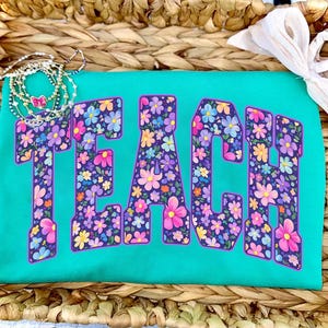 Op de afbeelding: Een turquoise t-shirt met het woord "TEACH" in grote, met bloemen gevulde letters. De letters zijn omrand met paars en gevuld met kleurrijke bloemen. Een ketting en een witte strik zijn ook zichtbaar.