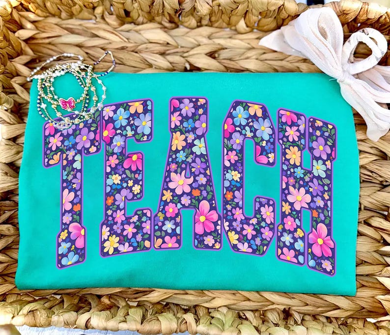 Op de afbeelding: Een turquoise t-shirt met het woord "TEACH" in grote, met bloemen gevulde letters. De letters zijn omrand met paars en gevuld met kleurrijke bloemen. Een ketting en een witte strik zijn ook zichtbaar.