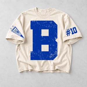 Könnte beinhalten: Ein cremefarbenes Kurzarm-T-Shirt mit einem großen, abgenutzten blauen Buchstaben B in der Mitte. Der rechte Ärmel zeigt eine blaue Wimpelgrafik mit dem Text BULLDOGS, während der linke Ärmel den blauen Text #10 aufweist.