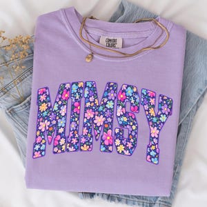 Op de afbeelding: Een lila sweatshirt met het woord "MIMSY" in grote, bloemrijke letters. Het sweatshirt is op een spijkerbroek gevouwen. Een gouden ketting met een kleine hanger ligt over het sweatshirt. Het merk "Comfort Colors" is zichtbaar.