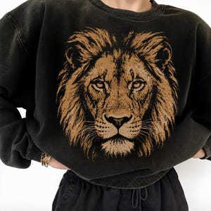 Könnte beinhalten: Ein anthrazitfarbenes Sweatshirt mit Rundhalsausschnitt und einem großen, detaillierten Grafikdruck eines Löwengesichts in Braun- und Beigetönen. Das Kleidungsstück hat einen gerippten Kragen, Bündchen und Saum sowie eine verwaschene Optik.