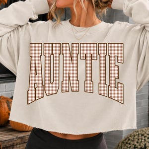 May include: A beige long-sleeve cropped sweatshirt featuring the word AUNTIE printed in a large, arched collegiate font. The letters have a brown and white gingham check pattern. The garment has a crew neckline and a raw, unfinished hemline.