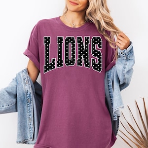 May include: A plum-colored short-sleeve t-shirt featuring the word LIONS in large, black, varsity-style block letters with a white polka dot pattern. The shirt has a crew neckline and a relaxed fit, layered under a light blue denim jacket.