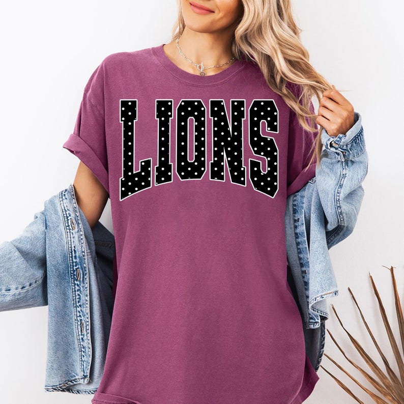 May include: A plum-colored short-sleeve t-shirt featuring the word LIONS in large, black, varsity-style block letters with a white polka dot pattern. The shirt has a crew neckline and a relaxed fit, layered under a light blue denim jacket.