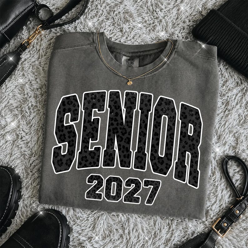 May include: A charcoal gray crewneck t-shirt featuring the word SENIOR in large, block-style letters with a black leopard print pattern and a white outline, followed by the number 2027 in the same leopard print style with a white outline.