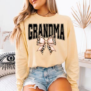 May include: A light yellow long-sleeve sweatshirt with the word "GRANDMA" in black leopard print letters. A bow with leopard print accents is centered below the word. The sweatshirt is paired with denim shorts.