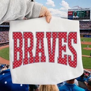 Op de afbeelding: Een wit t-shirt met het woord BRAVES in grote, dikke, rode blokletters met een wit stippenpatroon. De letters zijn omlijnd met een dunne donkerblauwe rand en zijn in een lichte boog in het midden van het kledingstuk geplaatst.