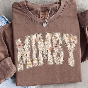 Op de afbeelding: Een bruine sweatshirt met het woord "MIMSY" in grote, bloemrijke letters. De letters zijn wit omlijnd en gevuld met een patroon van roze, oranje en gele bloemen en groene bladeren. Een zilveren ketting met een hanger is zichtbaar.