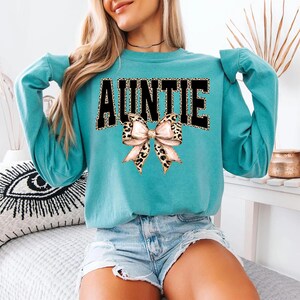 May include: A teal sweatshirt with the word "AUNTIE" in black block letters with a leopard print fill. A leopard print bow is centered below the text. The sweatshirt has long sleeves.