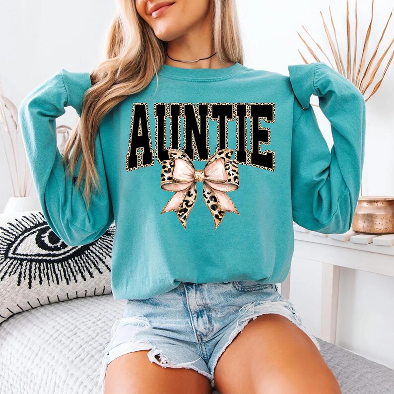 May include: A teal sweatshirt with the word "AUNTIE" in black block letters with a leopard print fill. A leopard print bow is centered below the text. The sweatshirt has long sleeves.