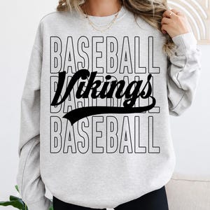 May include: A light gray crewneck sweatshirt featuring the word BASEBALL printed in a stacked, outlined font. Overlaid across the center is the word Vikings in a bold, black, cursive script with a swooping underline extending to the right.