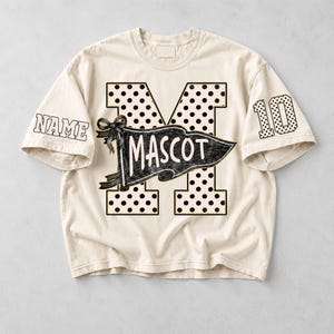May include: A cream-colored t-shirt featuring a large letter M with black polka dots. A black pennant graphic with the text MASCOT is layered over the letter. The left sleeve displays the text NAME, and the right sleeve features the number 10.