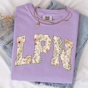 May include: A lilac Comfort Colors sweatshirt with the letters "LPN" in a floral design. The sweatshirt is folded on top of a pair of blue jeans. A gold necklace with a pendant is on top of the sweatshirt.