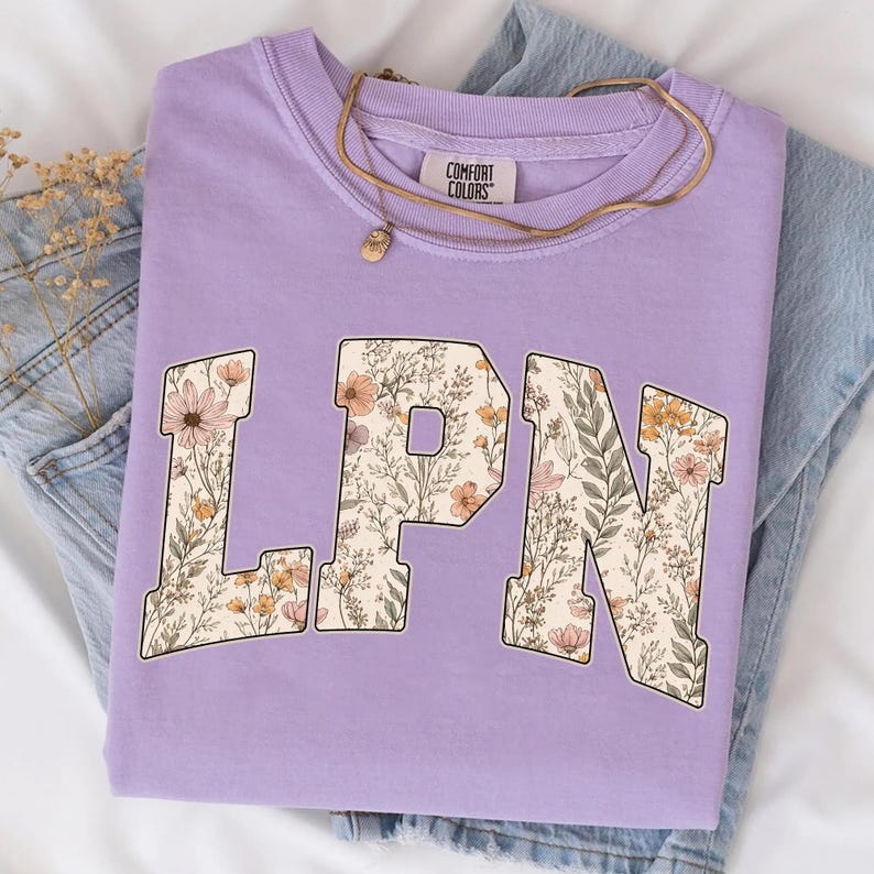 May include: A lilac Comfort Colors sweatshirt with the letters "LPN" in a floral design. The sweatshirt is folded on top of a pair of blue jeans. A gold necklace with a pendant is on top of the sweatshirt.