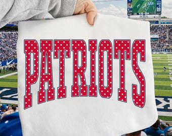 May include: A white fabric with the word "PATRIOTS" in large red letters with white polka dots. The letters have a blue outline. The background shows a football stadium with a scoreboard and spectators.