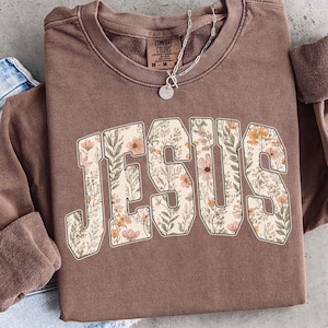 Peut inclure: Un sweat-shirt marron avec le mot "JESUS" en grandes lettres fleuries. Les lettres sont remplies d'un motif de fleurs et de feuilles roses, jaunes et vertes. Un collier argent&eacute; avec un pendentif est visible.