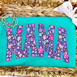 May include: A turquoise t-shirt with the word "MAMA" in large, floral-patterned letters. The flowers are in shades of pink, purple, yellow, and blue. The shirt is displayed on a woven basket.
