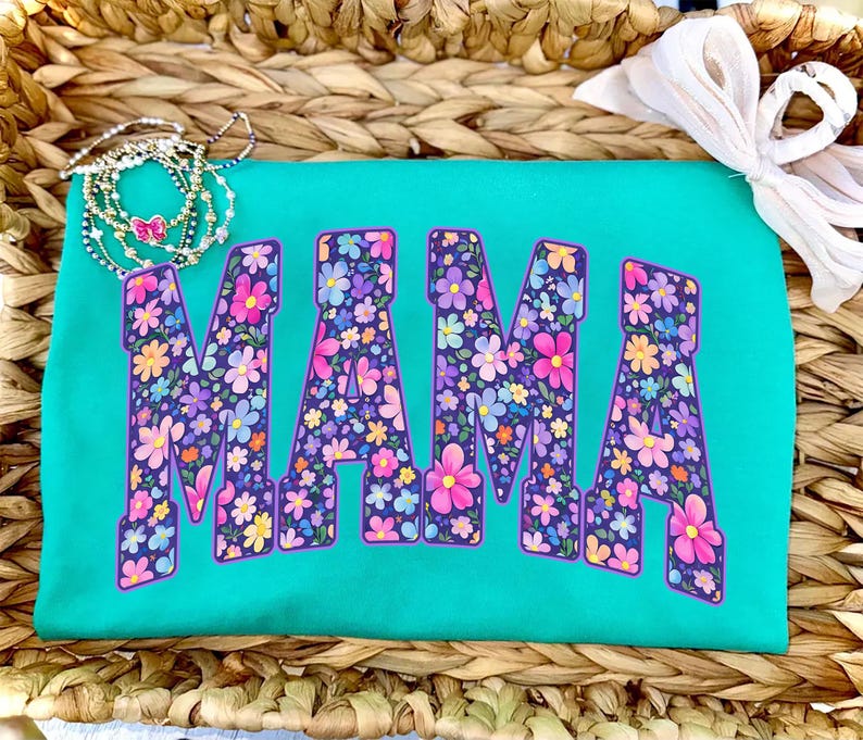May include: A turquoise t-shirt with the word "MAMA" in large, floral-patterned letters. The flowers are in shades of pink, purple, yellow, and blue. The shirt is displayed on a woven basket.