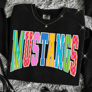 May include: A black crewneck sweatshirt featuring the word MUSTANGS printed in large, multicolored block letters. Each letter displays a unique pattern, including vertical stripes, red and orange checkers, pink polka dots, and blue triangles.