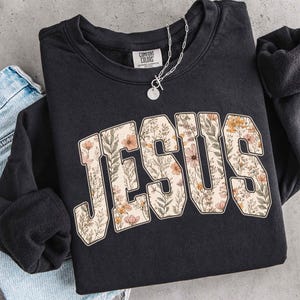 Peut inclure: Sweat-shirt noir Comfort Colors avec le mot "JESUS" dans un design floral. Les lettres sont remplies d'un fond cr&egrave;me et de diverses fleurs dans des tons de rose, d'orange et de vert. Un collier argent&eacute; avec un pendentif est drap&eacute; sur le sweat-shirt.