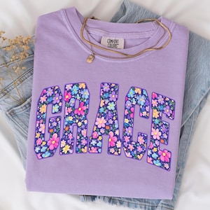 May include: A lilac Comfort Colors sweatshirt with the word "GRACE" in large, floral-filled letters. A gold necklace with a small pendant rests on the shirt. The sweatshirt is folded on top of a pair of blue jeans.