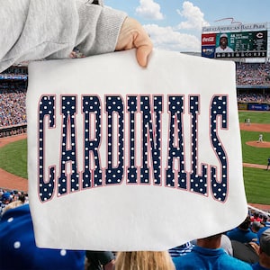 Może przedstawiać: Biały t-shirt z napisem CARDINALS wykonanym dużymi, ciemnoniebieskimi literami w białe kropki z cienkim czerwonym obrysem. Tekst jest wygięty w łuk na środku odzieży, kt&oacute;ra jest trzymana na tle stadionu baseballowego.
