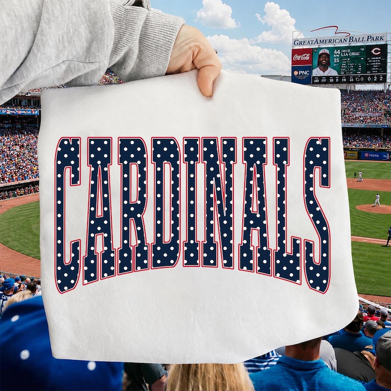 Może przedstawiać: Biały t-shirt z napisem CARDINALS wykonanym dużymi, ciemnoniebieskimi literami w białe kropki z cienkim czerwonym obrysem. Tekst jest wygięty w łuk na środku odzieży, kt&oacute;ra jest trzymana na tle stadionu baseballowego.