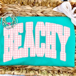 May include: A turquoise t-shirt with the word "BEACHY" in large, pink gingham letters. The shirt is accessorized with a stack of beaded bracelets and a white bow. The shirt is displayed on a woven basket.