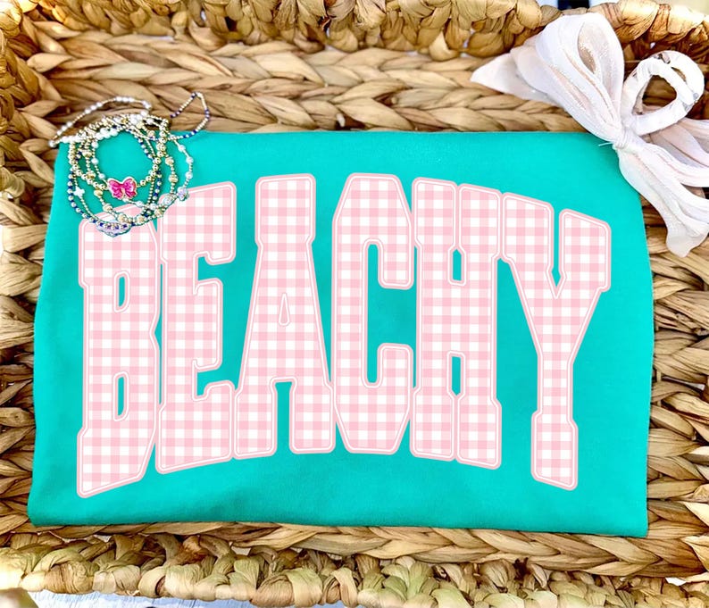 May include: A turquoise t-shirt with the word "BEACHY" in large, pink gingham letters. The shirt is accessorized with a stack of beaded bracelets and a white bow. The shirt is displayed on a woven basket.