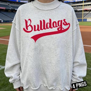 Könnte beinhalten: Ein hellgraues Sweatshirt mit dem Wort "Bulldogs" in roter Schrift auf der Vorderseite. Das Sweatshirt hat einen Rundhalsausschnitt und einen gewellten Saum. Der Hintergrund zeigt ein Baseballfeld.