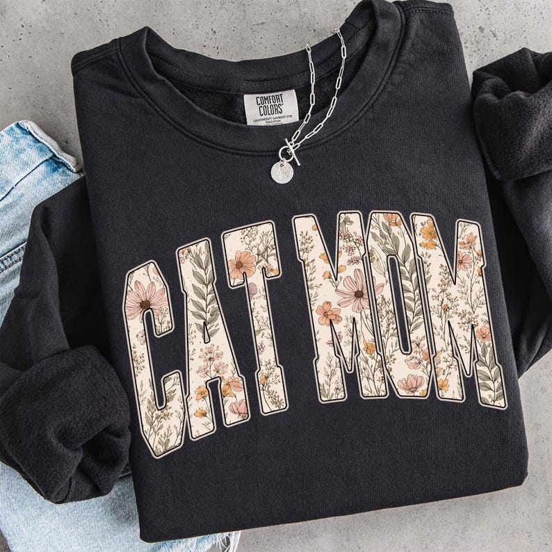 May include: Dark grey sweatshirt featuring the words "CAT MOM" in a floral design. The letters are filled with a pattern of pink, peach, and cream-coloured flowers and green leaves. A silver necklace with a pendant is on top.