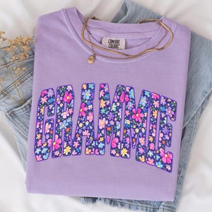 May include: A lilac Comfort Colors sweatshirt with the word "Grammie" in a floral design. The sweatshirt is folded on top of a pair of blue jeans. A gold necklace with a small pendant is draped across the shirt.