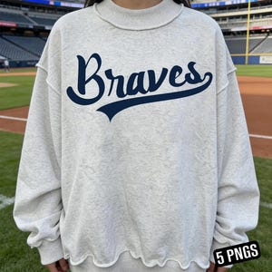 Op de afbeelding: Een lichtgrijs sweatshirt met het woord "Braves" in marineblauwe letters over de borst. De sweatshirt heeft een ronde hals en een golvende zoom. De achtergrond toont een honkbalveld.