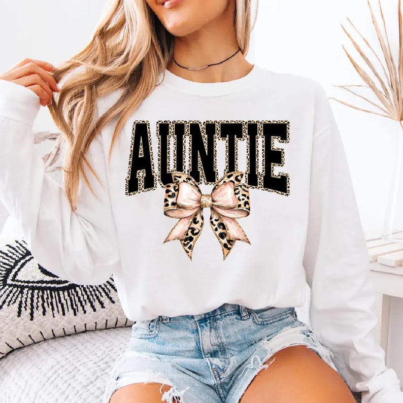 May include: White long-sleeve sweatshirt with the word "AUNTIE" in black block letters with a leopard print fill. A pink and leopard print bow is centered below the word. The sweatshirt is worn with denim shorts.