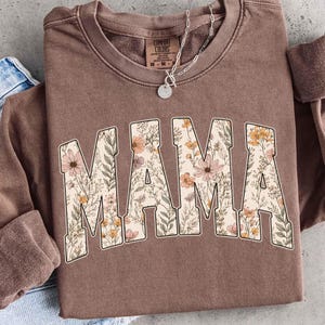 Könnte beinhalten: Ein braunes Sweatshirt mit dem Wort "MAMA" in großen Buchstaben mit floralem Muster auf der Vorderseite. Die Buchstaben sind mit einem Design aus rosa, pfirsichfarbenen und gelben Blumen und grünen Blättern gefüllt. Eine silberne Halskette mit einem kleinen Anhänger ist sichtbar.