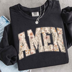 May include: Black sweatshirt with the word "AMEN" in a floral design. The letters are filled with a pattern of flowers in shades of pink, yellow, and green. A silver necklace with a pendant is draped over the sweatshirt.