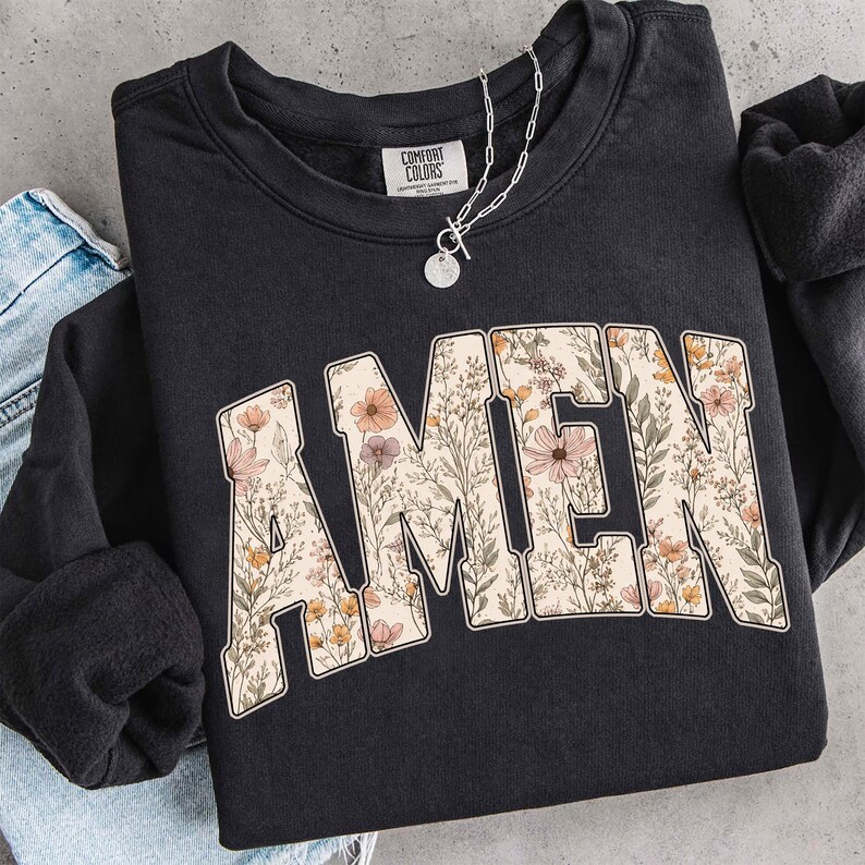 May include: Black sweatshirt with the word "AMEN" in a floral design. The letters are filled with a pattern of flowers in shades of pink, yellow, and green. A silver necklace with a pendant is draped over the sweatshirt.