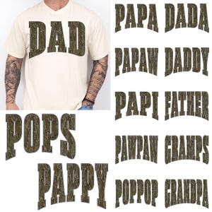 May include: A cream-coloured t-shirt with the word "DAD" in a textured, camouflage-style font. Additional text options include "PAPA", "DADA", "PAPAW", "DADDY", "POPS", "PAPPY", "PAPI", "FATHER", "PANPAW", "GRAMPS", "POPOOP", and "GRANDPA".