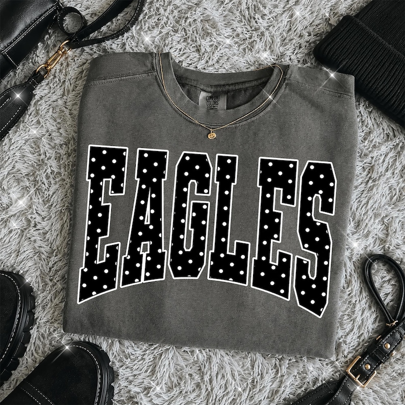 May include: A charcoal gray crewneck sweatshirt featuring the word EAGLES in large, bold, black block letters with a white polka dot pattern. The garment has a ribbed neckline and a visible brand tag inside the collar, presented in a flat lay.