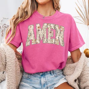 May include: A pink t-shirt with the word "AMEN" in a floral design across the front. The letters are filled with a variety of flowers in shades of beige, orange, and green. The shirt has short sleeves and a crew neck.