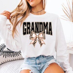 May include: White long-sleeve sweatshirt with the word "GRANDMA" in black block letters with a leopard print fill. A bow with a leopard print pattern is below the word. The sweatshirt is a casual style.
