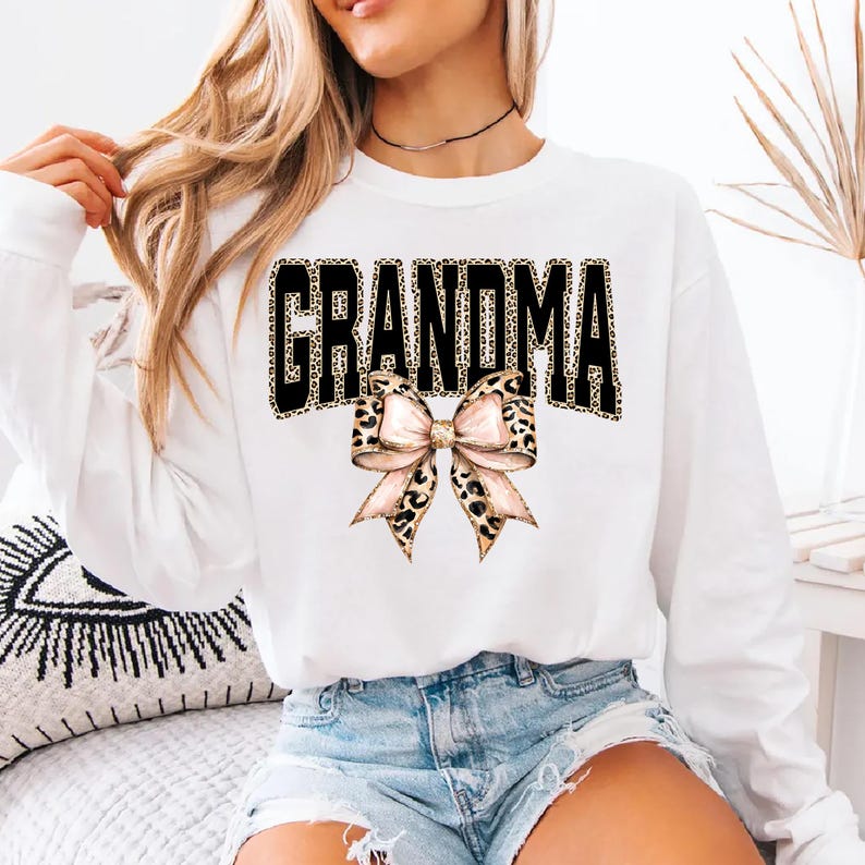 May include: White long-sleeve sweatshirt with the word "GRANDMA" in black block letters with a leopard print fill. A bow with a leopard print pattern is below the word. The sweatshirt is a casual style.