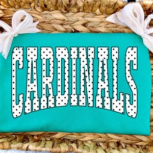 Może przedstawiać: Jaskrawo turkusowy t-shirt z napisem CARDINALS wykonanym dużymi, białymi literami blokowymi w czarne kropki. Odzież jest ozdobiona dwiema białymi kokardami z materiału przymocowanymi do g&oacute;rnych rog&oacute;w koszulki.
