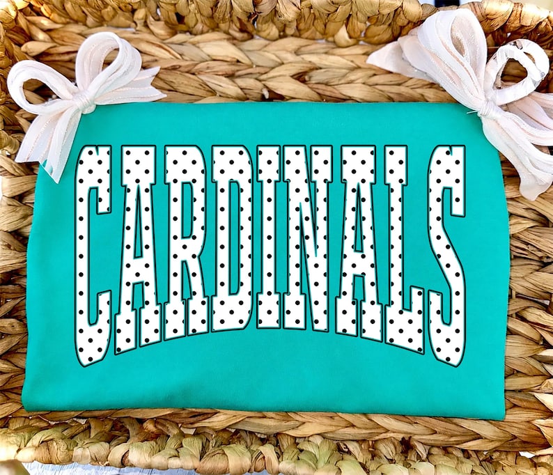 Może przedstawiać: Jaskrawo turkusowy t-shirt z napisem CARDINALS wykonanym dużymi, białymi literami blokowymi w czarne kropki. Odzież jest ozdobiona dwiema białymi kokardami z materiału przymocowanymi do g&oacute;rnych rog&oacute;w koszulki.