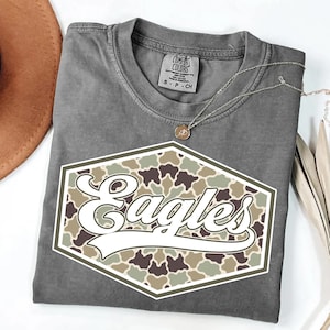 May include: A gray Comfort Colors t-shirt with a white and camouflage "Eagles" graphic. The graphic is in a hexagon shape. A silver necklace with a small pendant is on the shirt.