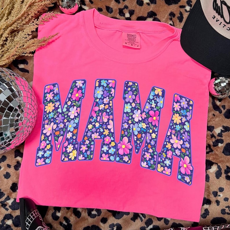 May include: A bright pink t-shirt with the word "MAMA" in large, floral-filled letters. The flowers are in shades of pink, purple, yellow, and blue. The shirt has a crew neck and short sleeves. A black baseball cap is visible in the upper right corner.