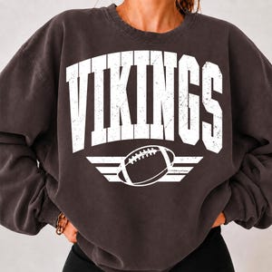 Op de afbeelding: Een antracietgrijze sweater met ronde hals en het woord VIKINGS in grote, verweerde witte blokletters op de borst. Onder de tekst staat een afbeelding van een football gecentreerd tussen twee horizontale witte lijnen.