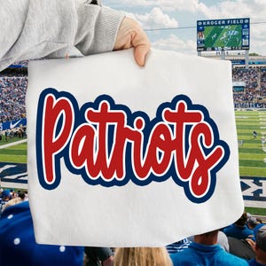 May include: White rectangular sign with the word "Patriots" in red script with a white outline and a navy blue border. The sign is held up in front of a football stadium.