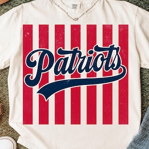 May include: An off-white short-sleeve t-shirt featuring a large rectangular graphic on the front. The graphic consists of vertical red and white stripes with the word Patriots written in a dark blue script font across the center.