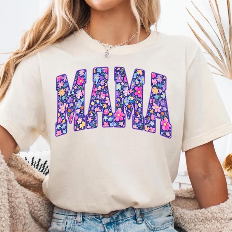 May include: Cream-colored t-shirt with the word "MAMA" in large, purple, floral-filled letters. The flowers are in shades of pink, blue, yellow, and purple. The shirt has short sleeves and a relaxed fit.